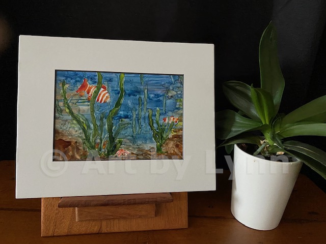 Framed watercolor of underwater scene with clownfish and seaweed, next to a house plant.